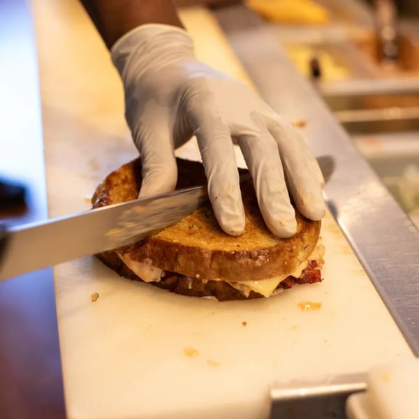 A gloved hand slicing a grilled cheese sandwich on a white cutting board at JD's Cafe.